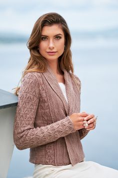 a woman wearing a cardigan and sitting on a ledge next to the water with her hands in her pockets