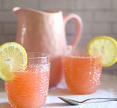 two glasses filled with orange liquid and lemon wedges next to a pitcher on a table