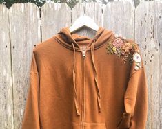 a brown hoodie with flowers on it is hanging against a wooden fence and has a white hanger in front of it