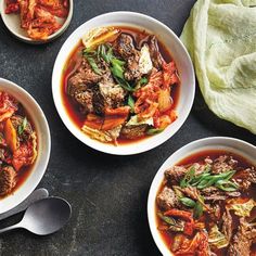 three bowls of soup with meat and vegetables in them on a black surface next to two spoons