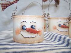 three pots with faces painted on them sitting on a striped table cloth, one has an orange nose and the other has blue eyes