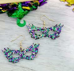 pair of purple and green mardi gras mask earrings on white wooden table next to confetti