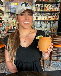 a woman holding a drink in her right hand and smiling at the camera while wearing a hat