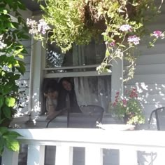 two women are looking out the window in front of their house with flowers on the porch