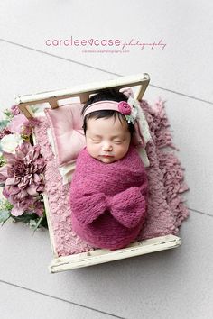 a baby wrapped in a pink blanket laying on top of a wooden crate next to flowers