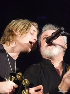 two men singing into microphones on stage with one holding his head in the other's hand