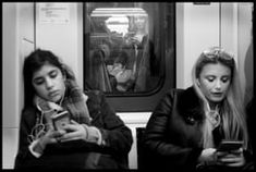 two women sitting on a subway looking at their cell phones while one looks at her phone