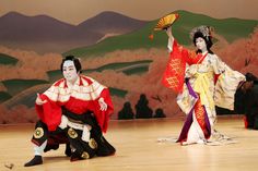 two people dressed in geisha outfits on stage with one holding a fan and the other sitting down