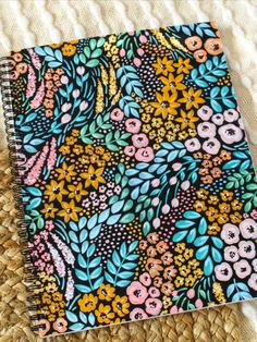 a notebook with colorful flowers on it sitting on top of a woven table cloth next to a straw bag