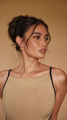 a woman with dark hair wearing a tan top and gold earrings, standing in front of a beige wall