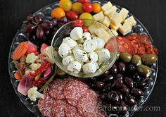 an assortment of meats and cheeses on a platter with olives, tomatoes, grapes, bread