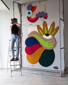 a woman standing on a ladder in front of a wall with colorful art hanging from it