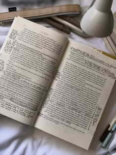 an open book sitting on top of a bed next to a lamp and some pens