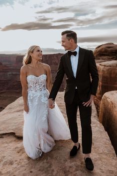 a bride and groom holding hands while standing on top of a cliff in the desert