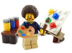 a lego man holding a paintbrush and palette in front of an easel with paints on it