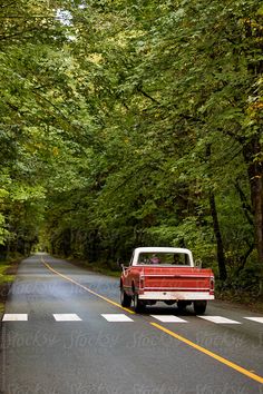 an old red pick up truck driving down the road in front of some green trees