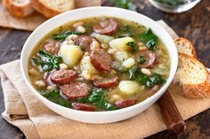 a bowl of soup with sausage, potatoes and spinach on a napkin next to bread