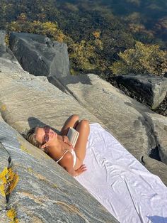 a woman laying on top of a rock next to a body of water with an open book in her hand