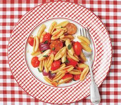 a white plate topped with pasta covered in tomatoes and sausage next to a fork on a checkered table cloth