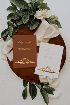 the wedding stationery is laid out on a wooden platter with flowers and greenery