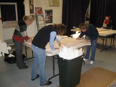 three people standing around a table with boxes on it and one person holding a paper