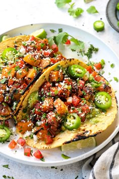 two tacos on a white plate topped with tomatoes, peppers and seasoning sprinkles