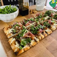 a wooden cutting board topped with slices of pizza next to a bowl of greens and nuts