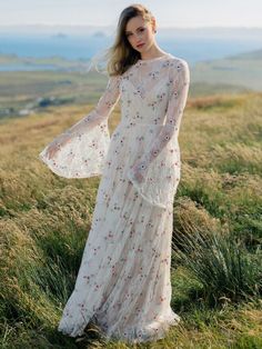 a woman standing on top of a grass covered hillside wearing a white dress with red flowers