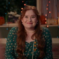 a woman with long red hair wearing a green dress and pearls on her necklace smiles at the camera