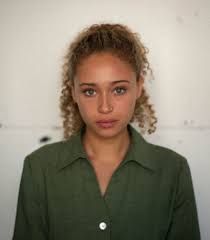 a woman with curly hair and green shirt looking at the camera while standing in front of a white wall