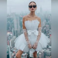 a woman in a short white dress and gloves standing on top of a building with her hands behind her back