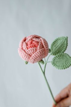 a pink crocheted rose being held by someone's hand with green leaves