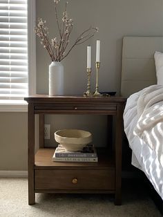 a nightstand with a bowl and candles on it in front of a bed, next to a window