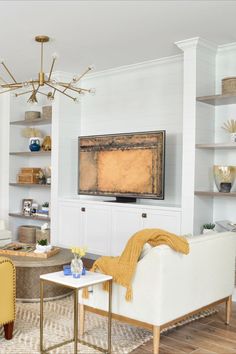 This image shows a modern living room design with a gold chandelier, a beige area rug, a white shiplap wall with built-ins, a cream loveseat, a gold side table, and modern decor items. Design by One Coast Design, located in Summerville, SC. Cream Loveseat, Bronze Coffee Table, Coastal Decorating Living Room, Modern Coastal Decor, Coastal Living Room, Decorating Shelves, Soft Beige