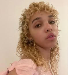 a close up of a person with curly hair wearing a pink shirt and earrings looking at the camera