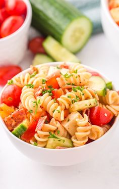 a white bowl filled with pasta salad next to cucumbers and tomatoes