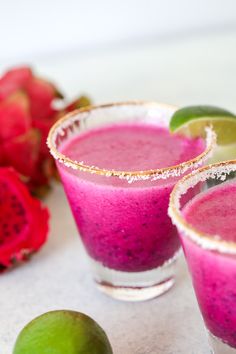 two glasses filled with pink and green smoothie next to some fruit on the table