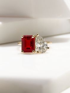 an engagement ring with two pear shaped diamonds and a red stone in the center, sitting on a white surface