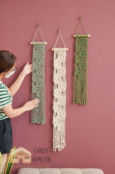 a woman standing in front of three macrame wall hangings on a pink wall