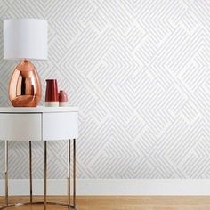 a white table with a lamp and vase on it in front of a patterned wall