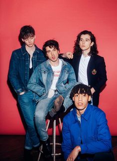 three young men sitting on stools in front of a red wall, one wearing a denim jacket