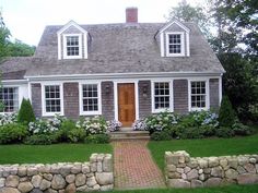 a small gray house surrounded by green grass and flowers with a brick walkway leading to the front door