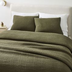 a bed with green bedspread and pillows in a white walled room next to a night stand