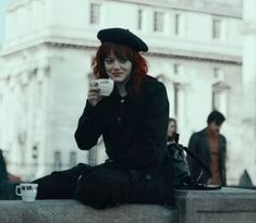 a woman with red hair sitting on a ledge drinking from a cup