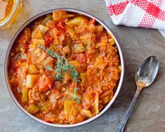 a bowl filled with pasta and vegetables on top of a wooden table next to a spoon