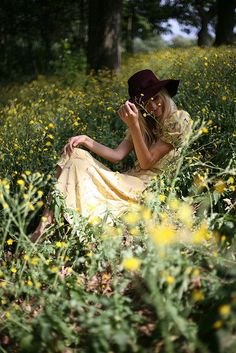 a woman in a yellow dress and hat sitting on the ground surrounded by wildflowers