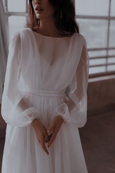 a woman standing in front of a window wearing a white dress and holding her hands together