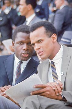 two men sitting next to each other in suits and ties looking at something on a piece of paper