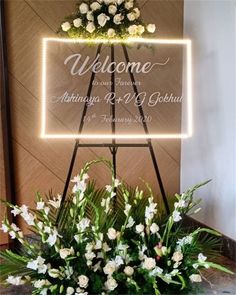 a welcome sign with flowers and greenery in front of it on a table next to a planter