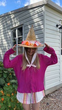 a woman wearing a straw hat standing in front of a house with her hands behind her head